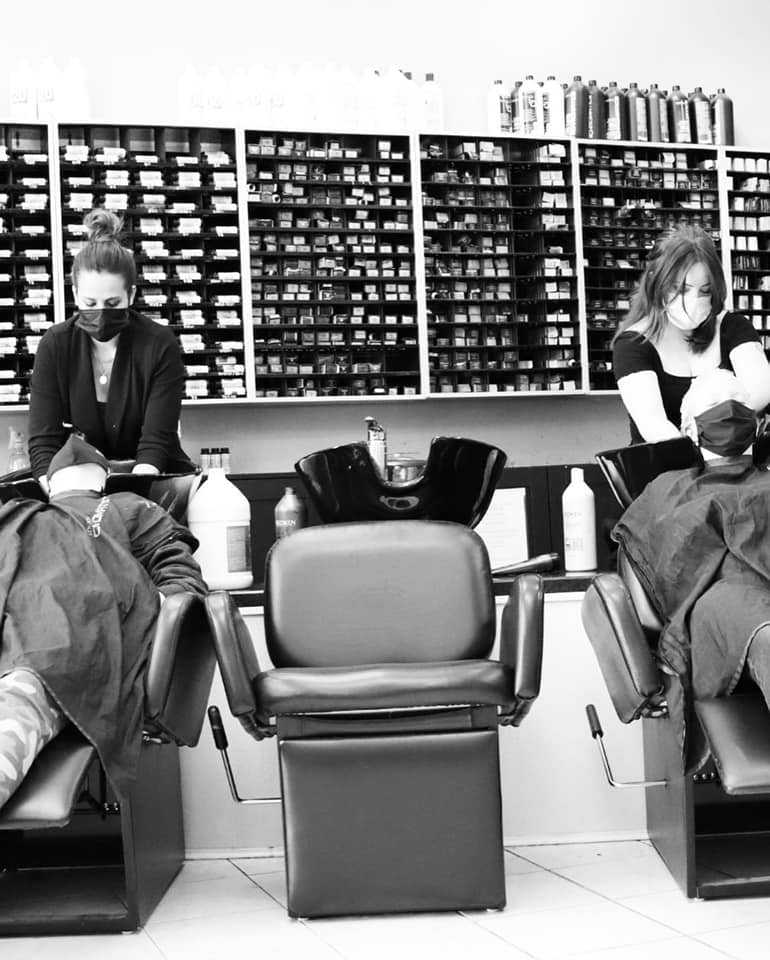 Two people sitting in salon chairs in front of hair product shelves.