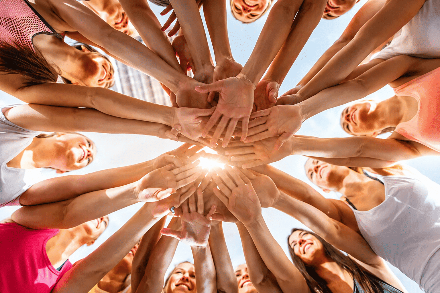 Group of people in a circle, stacking hands together under a clear blue sky and sunlight.