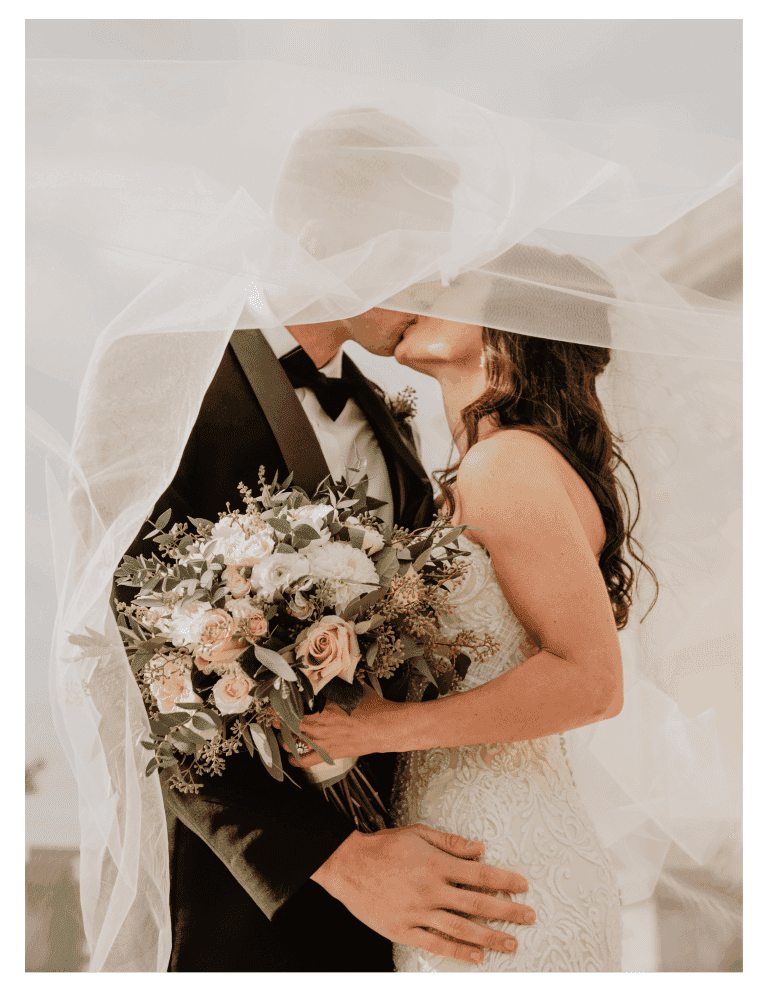 Bride and groom kissing under veil, holding a bouquet.