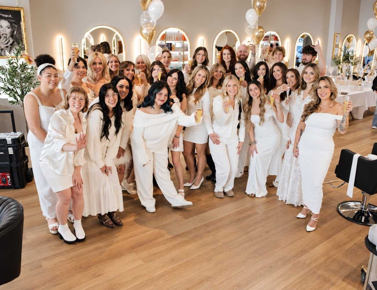 Group of people in white clothing celebrating indoors with drinks and balloons.